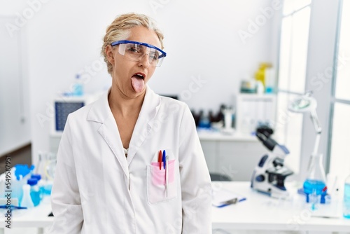 Middle age blonde woman working at scientist laboratory sticking tongue out happy with funny expression. emotion concept.