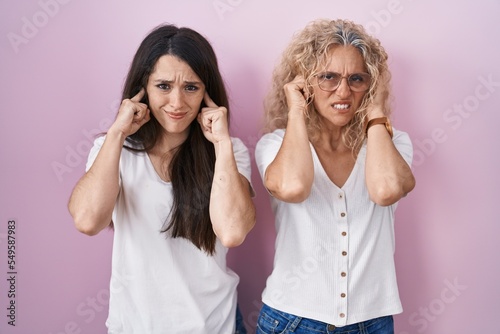 Mother and daughter standing together over pink background covering ears with fingers with annoyed expression for the noise of loud music. deaf concept.