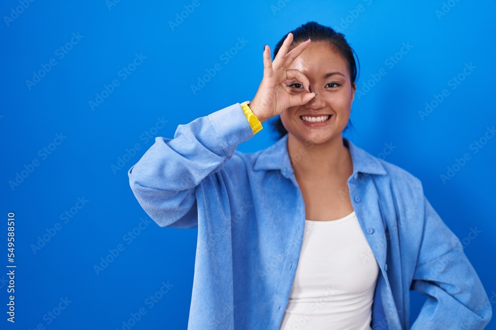 Asian young woman standing over blue background doing ok gesture with hand smiling, eye looking through fingers with happy face. Asian young woman standing over blue background doing ok gesture with hand smiling, eye looking through fingers with happy face.