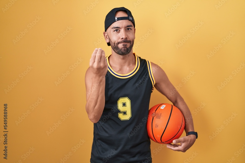 Middle age bald man holding basketball ball over yellow background doing italian gesture with hand and fingers confident expression Middle age bald man holding basketball ball over yellow background doing italian gesture with hand and fingers confident expression