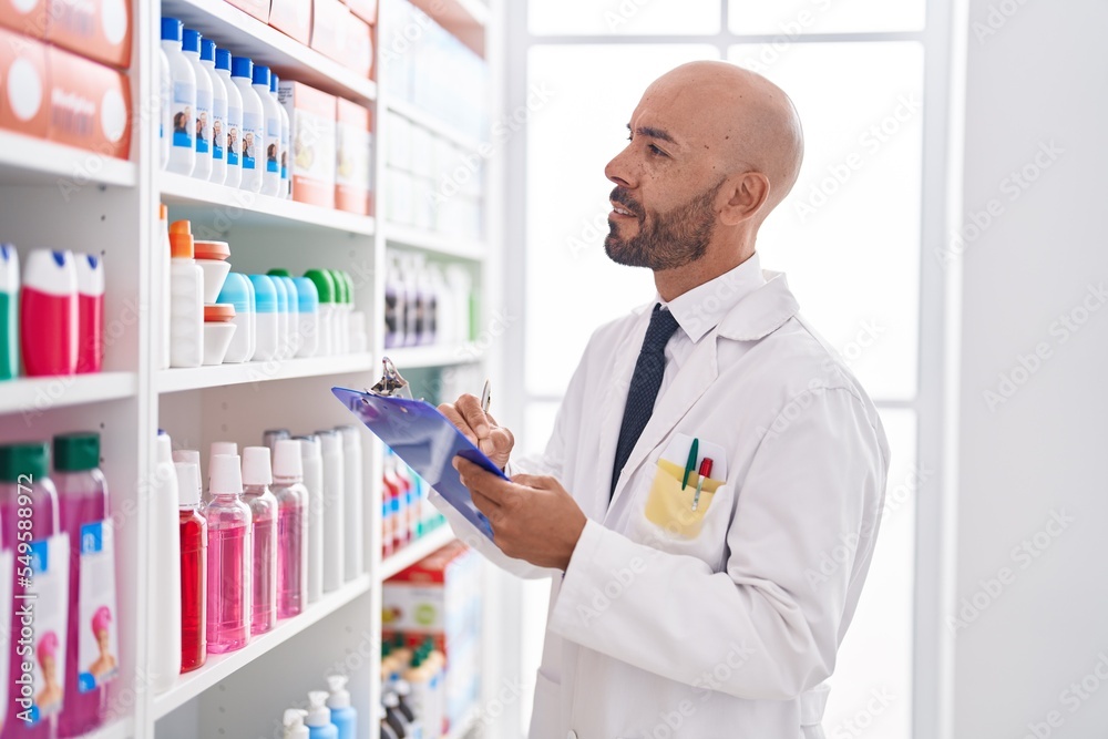 Young bald man pharmacist writing on document at pharmacy