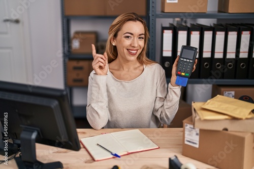 Hispanic woman working at small business ecommerce holding credit card and dataphone surprised with an idea or question pointing finger with happy face, number one
