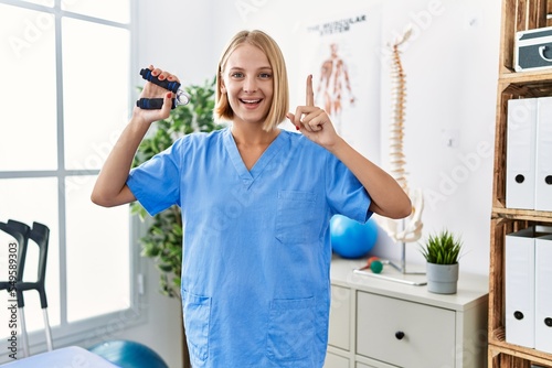 Young caucasian physiotherapist woman holding hand grip to train muscle smiling with an idea or question pointing finger with happy face  number one