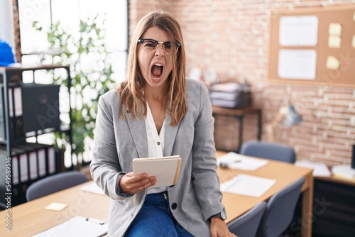 Young hispanic woman working at the office wearing glasses angry and mad screaming frustrated and furious, shouting with anger. rage and aggressive concept.