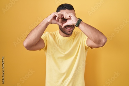 Young hispanic man standing over yellow background doing heart shape with hand and fingers smiling looking through sign © Krakenimages.com
