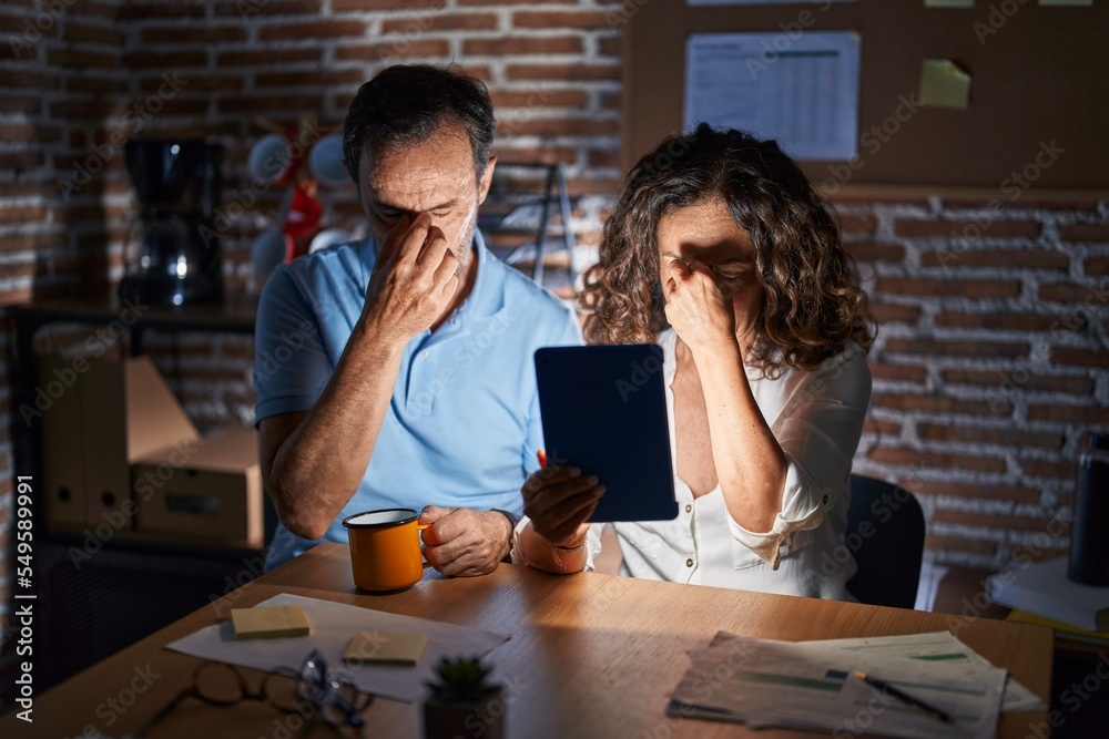 Middle age hispanic couple using touchpad sitting on the table at night tired rubbing nose and eyes feeling fatigue and headache. stress and frustration concept. Middle age hispanic couple using touchpad sitting on the table at night tired rubbing nose and eyes feeling fatigue and headache. stress and frustration concept.