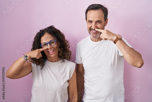 Middle age hispanic couple together over pink background pointing with hand finger to face and nose, smiling cheerful. beauty concept