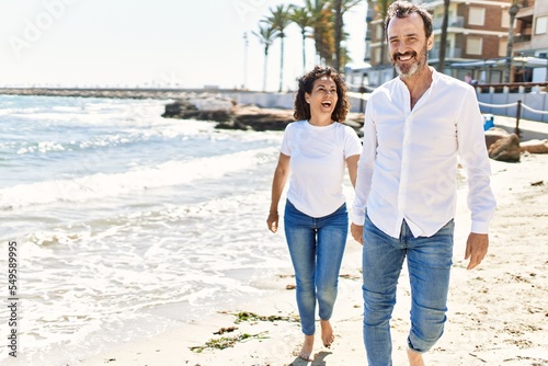 Middle age hispanic couple smiling happy walking with hands together at the beach.