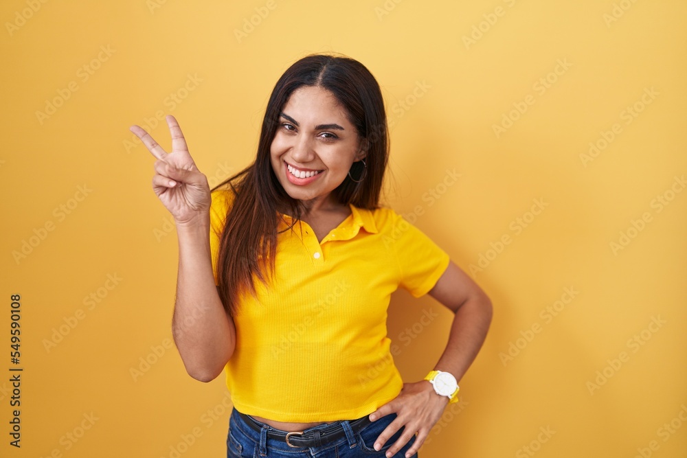 Young arab woman standing over yellow background smiling looking to the camera showing fingers doing victory sign. number two. Young arab woman standing over yellow background smiling looking to the camera showing fingers doing victory sign. number two.