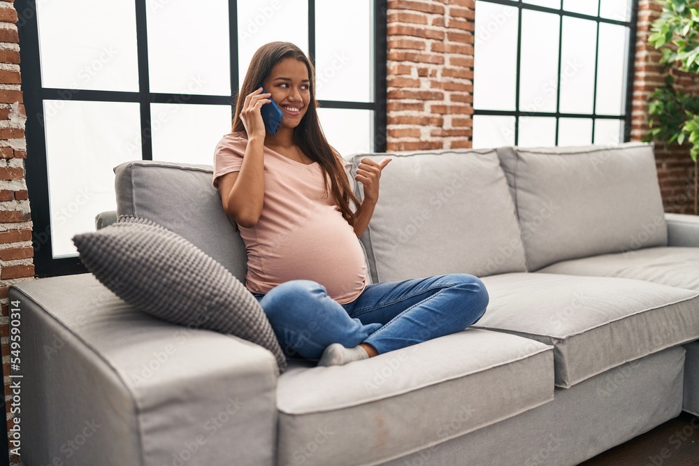 Fototapeta premium Young pregnant woman having conversation speaking on the smartphone pointing thumb up to the side smiling happy with open mouth