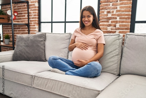 Young latin woman pregnant touching belly sitting on sofa at home