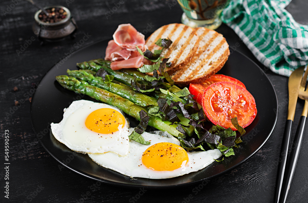 Breakfast. Fried egg, bread toast, green asparagus, tomatoes and jamon on black plate. Breakfast. Fried egg, bread toast, green asparagus, tomatoes and jamon on black plate.
