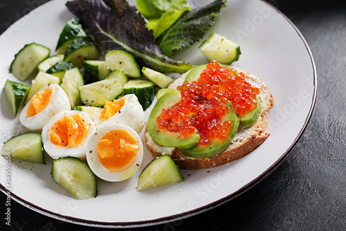 Breakfast. Healthy open sandwich on  toast with avocado and red caviar, boiled eggs, cucumber salad on white plate. Healthy protein food.
