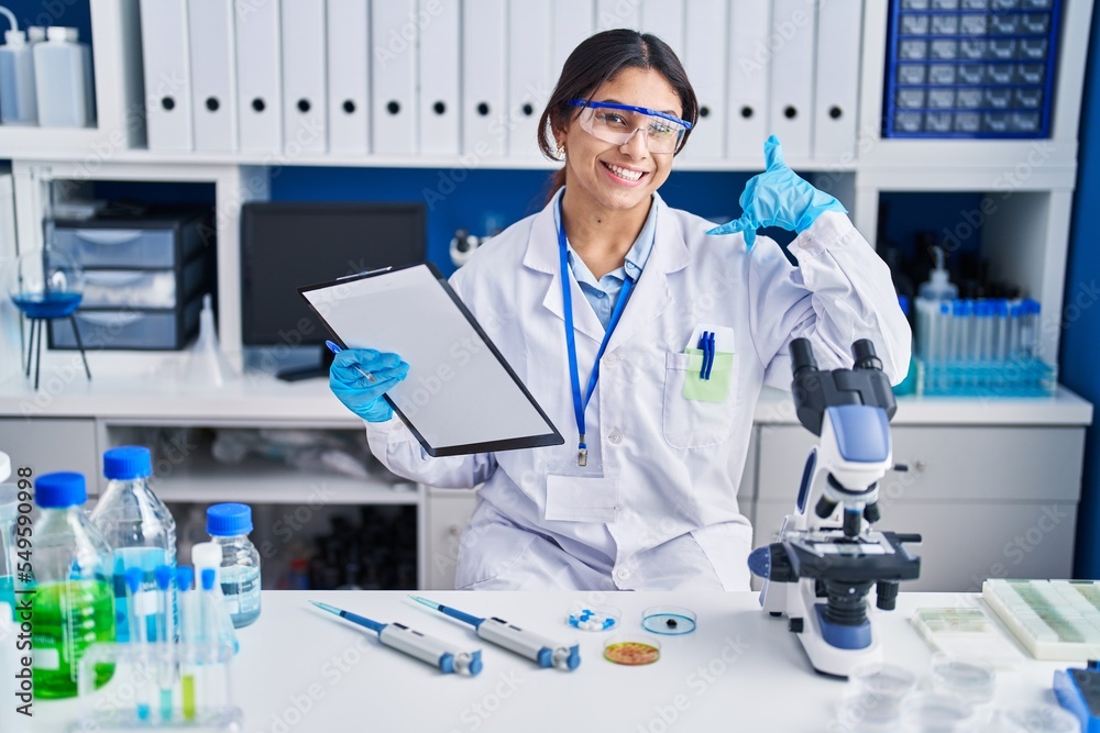 Hispanic young woman working at scientist laboratory smiling doing phone gesture with hand and fingers like talking on the telephone. communicating concepts.