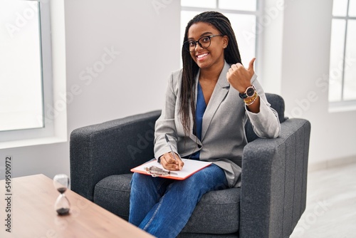 Young african american with braids working at consultation office pointing to the back behind with hand and thumbs up  smiling confident