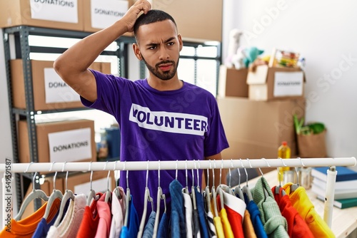 African american man wearing volunteer t shirt at donations stand confuse and wonder about question. uncertain with doubt, thinking with hand on head. pensive concept.