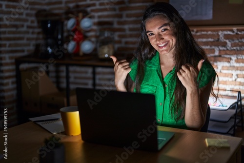 Young teenager girl working at the office at night success sign doing positive gesture with hand, thumbs up smiling and happy. cheerful expression and winner gesture.