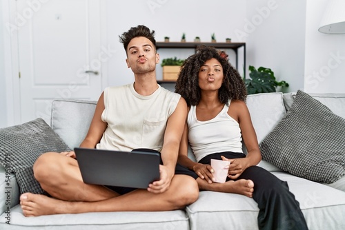 Young interracial couple using laptop at home sitting on the sofa looking at the camera blowing a kiss on air being lovely and sexy. love expression.