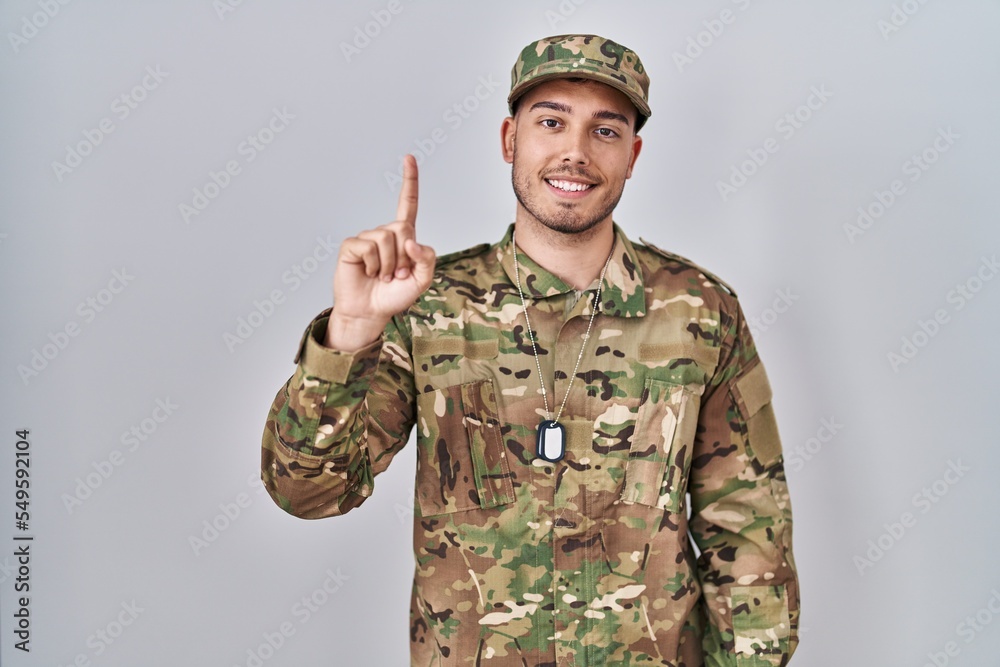 Young hispanic man wearing camouflage army uniform showing and pointing up with finger number one while smiling confident and happy. Young hispanic man wearing camouflage army uniform showing and pointing up with finger number one while smiling confident and happy.