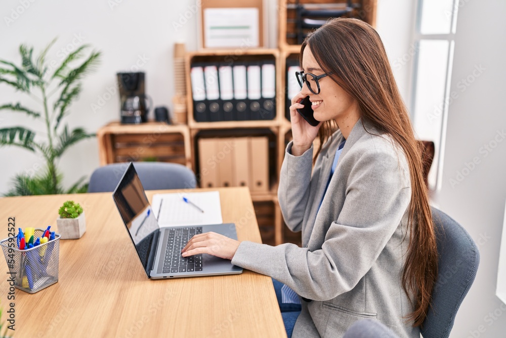 Young beautiful hispanic woman business worker using laptop talking on smartphone at office Young beautiful hispanic woman business worker using laptop talking on smartphone at office