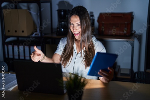 Young brunette woman working at the office at night approving doing positive gesture with hand, thumbs up smiling and happy for success. winner gesture.
