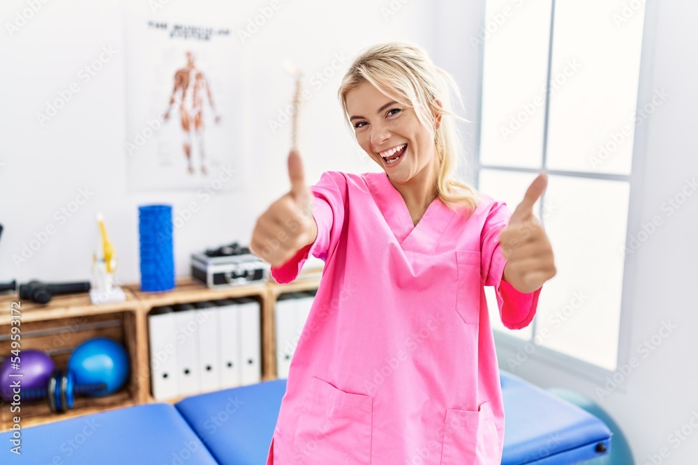 Young caucasian woman working at pain recovery clinic approving doing positive gesture with hand, thumbs up smiling and happy for success. winner gesture.