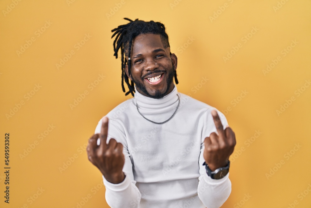 African man with dreadlocks wearing turtleneck sweater over yellow background showing middle finger doing fuck you bad expression, provocation and rude attitude. screaming excited African man with dreadlocks wearing turtleneck sweater over yellow background showing middle finger doing fuck you bad expression, provocation and rude attitude. screaming excited