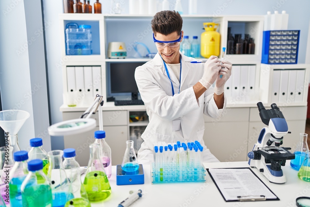 Fototapeta premium Young hispanic man wearing scientist uniform writing on test tube at laboratory