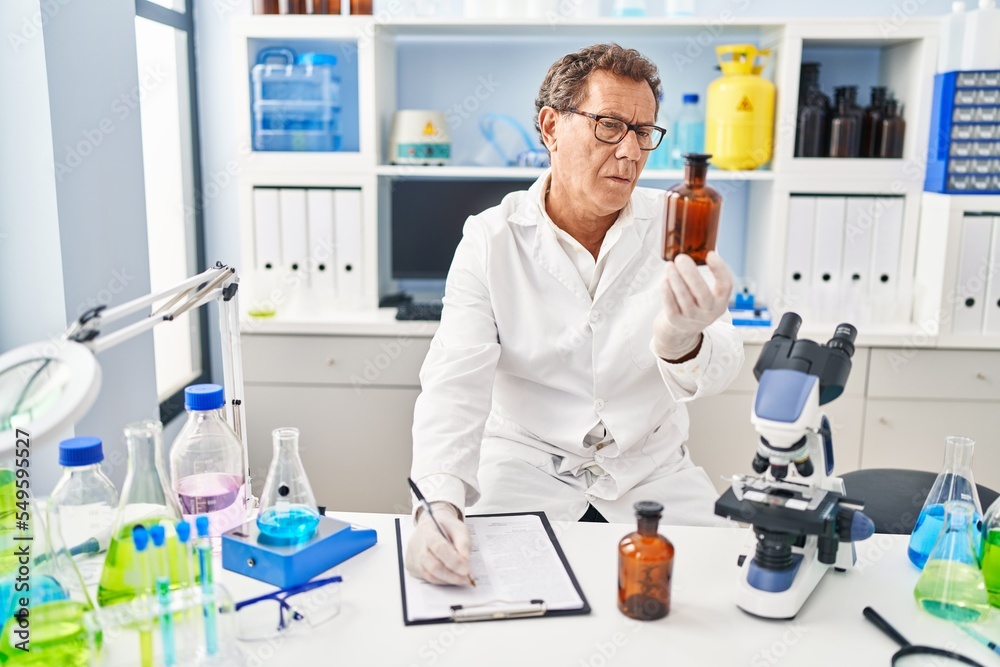 Middle age man wearing scientist uniform holding bottle writing on clipboard at laboratory Middle age man wearing scientist uniform holding bottle writing on clipboard at laboratory