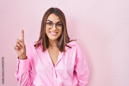 Young hispanic woman wearing glasses standing over pink background showing and pointing up with finger number one while smiling confident and happy.