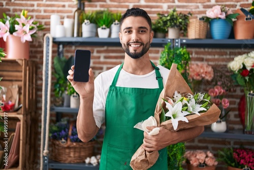 Hispanic young man working at florist shop showing smartphone screen smiling with a happy and cool smile on face. showing teeth.