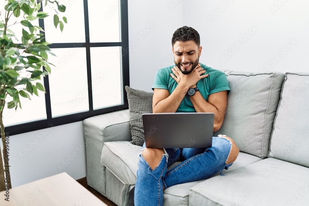 Young handsome man with beard using computer laptop sitting on the sofa at home shouting and suffocate because painful strangle. health problem. asphyxiate and suicide concept.