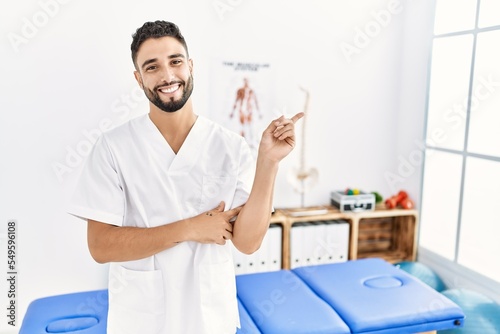 Young handsome man with beard working at pain recovery clinic with a big smile on face, pointing with hand and finger to the side looking at the camera.