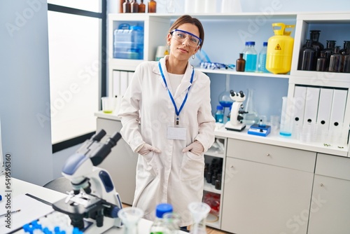 Young hispanic woman wearing scientist uniform standing at laboratory