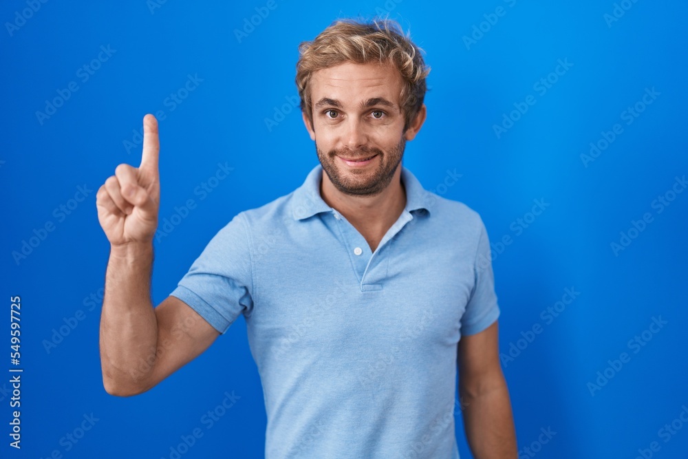Caucasian man standing over blue background showing and pointing up with finger number one while smiling confident and happy. Caucasian man standing over blue background showing and pointing up with finger number one while smiling confident and happy.