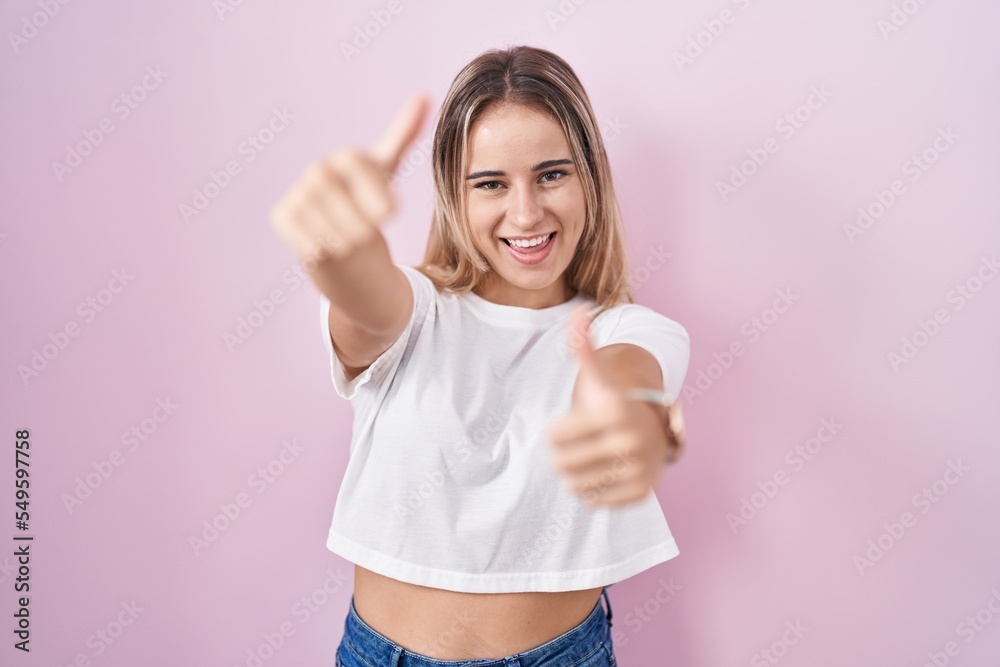 Young blonde woman standing over pink background approving doing positive gesture with hand, thumbs up smiling and happy for success. winner gesture. Young blonde woman standing over pink background approving doing positive gesture with hand, thumbs up smiling and happy for success. winner gesture.