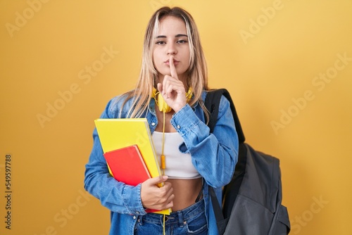 Young blonde woman wearing student backpack and holding books asking to be quiet with finger on lips. silence and secret concept.