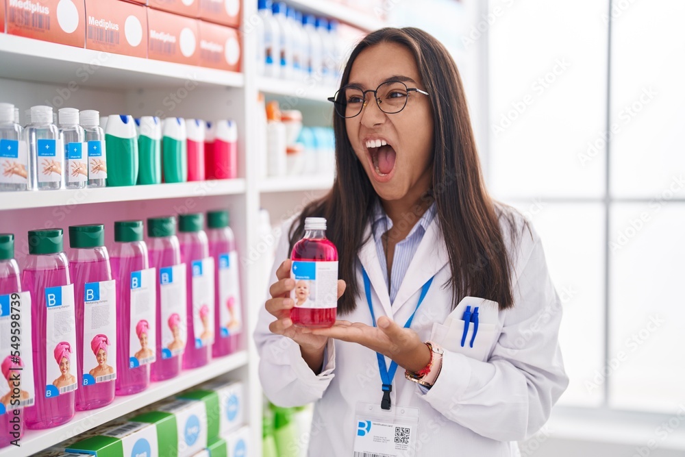 Young hispanic woman working at pharmacy drugstore holding syrup angry and mad screaming frustrated and furious, shouting with anger. rage and aggressive concept. Young hispanic woman working at pharmacy drugstore holding syrup angry and mad screaming frustrated and furious, shouting with anger. rage and aggressive concept.