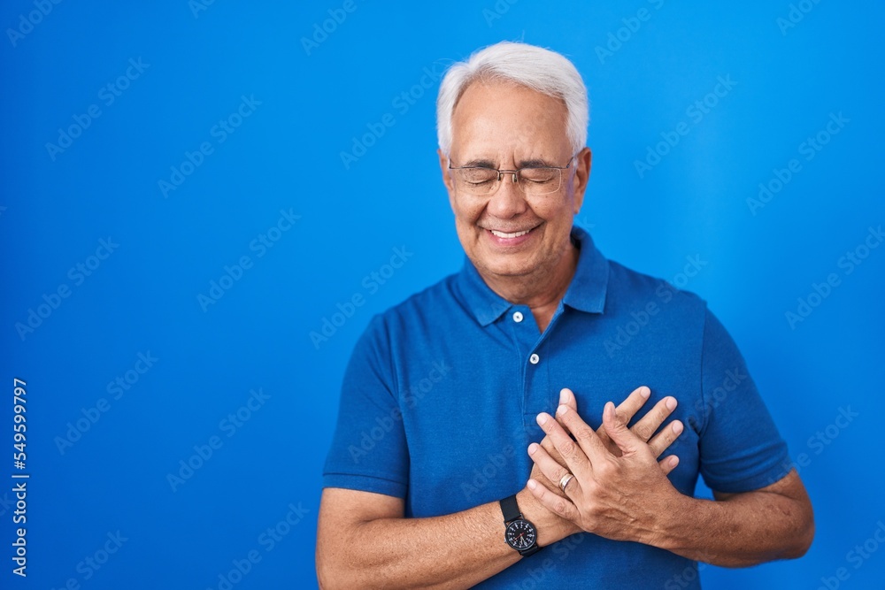 Middle age man with grey hair standing over blue background smiling with hands on chest with closed eyes and grateful gesture on face. health concept. Middle age man with grey hair standing over blue background smiling with hands on chest with closed eyes and grateful gesture on face. health concept.