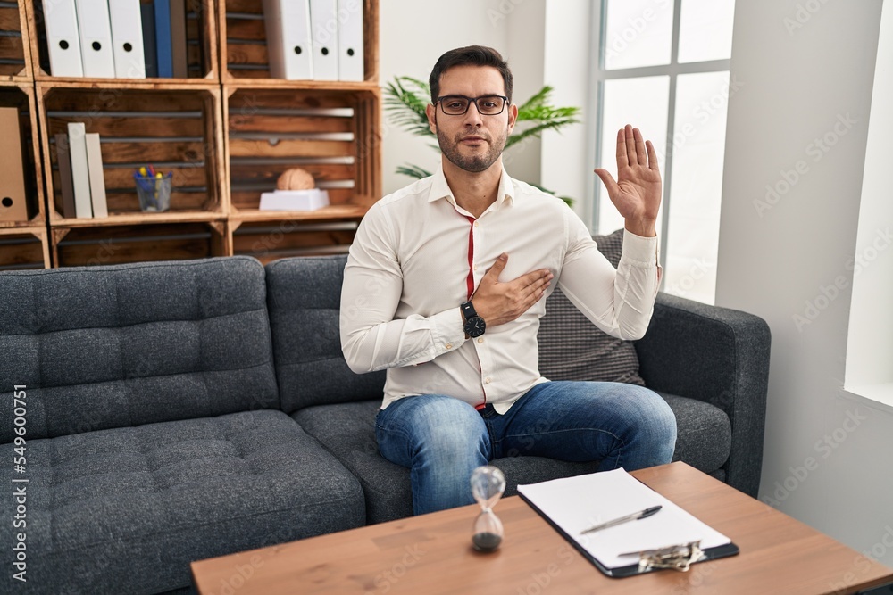 Young hispanic man with beard working at consultation office swearing with hand on chest and open palm, making a loyalty promise oath Young hispanic man with beard working at consultation office swearing with hand on chest and open palm, making a loyalty promise oath