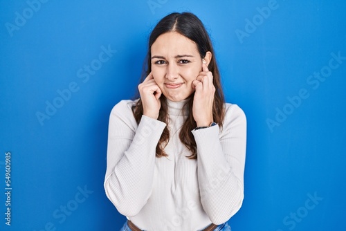 Young hispanic woman standing over blue background covering ears with fingers with annoyed expression for the noise of loud music. deaf concept.