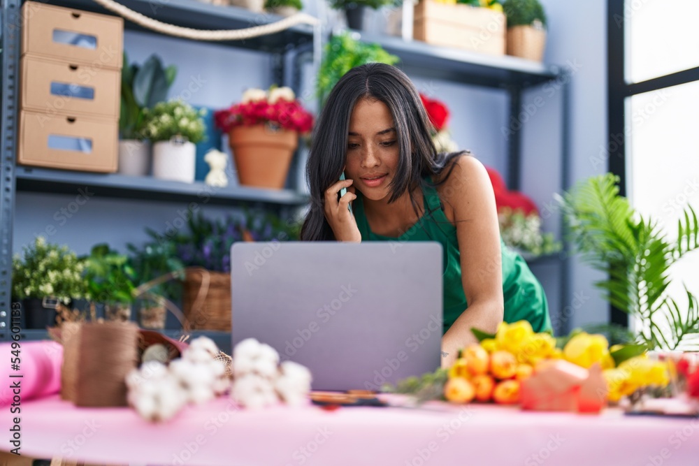 Young hispanic woman florist using laptop talking on smartphone at florist shop