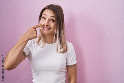 Blonde caucasian woman standing over pink background pointing with hand finger to face and nose, smiling cheerful. beauty concept