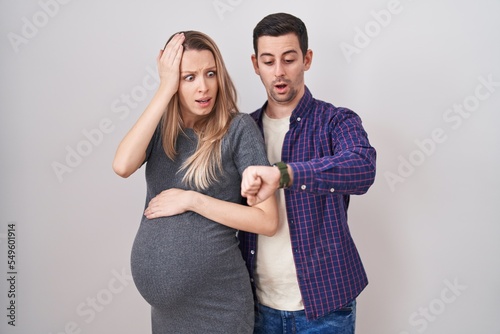 Young couple expecting a baby standing over white background looking at the watch time worried, afraid of getting late