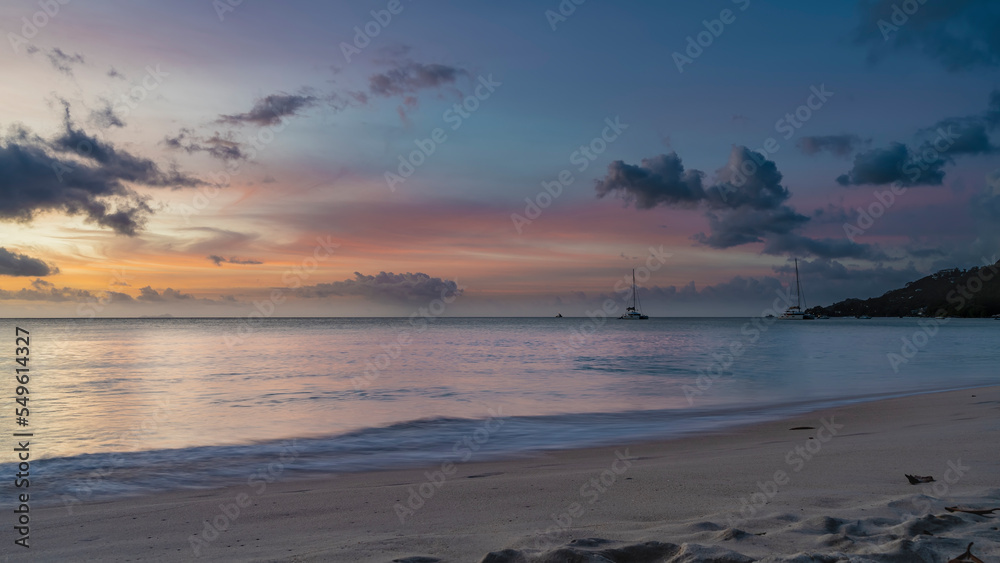 Romantic evening on a tropical beach. Blue clouds in the sky, highlighted in pink, scarlet, orange. Yachts and a hill on the horizon. Foam of waves on the sand. Long exposure. Seychelles. Mahe.