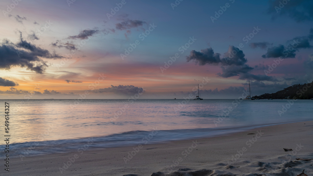 Romantic evening on a tropical beach. Blue clouds in the sky, highlighted in pink, scarlet, orange. Yachts and a hill on the horizon. Foam of waves on the sand. Long exposure. Seychelles. Mahe. Romantic evening on a tropical beach. Blue clouds in the sky, highlighted in pink, scarlet, orange. Yachts and a hill on the horizon. Foam of waves on the sand. Long exposure. Seychelles. Mahe.