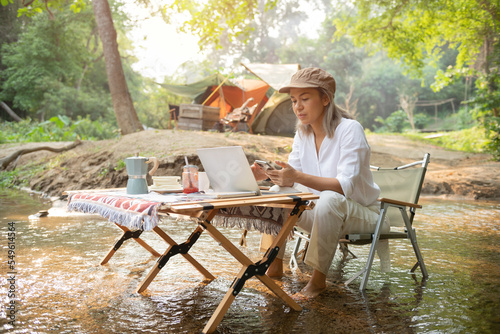 Asian woman travel and camping alone reading a book and using laptop while relaxing on the deck chairs in the river at natural park in Thailand. Recreation and journey outdoor activity lifestyle. © nareekarn