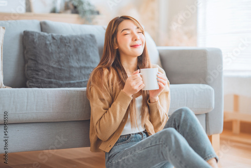 Joyful young asian female enjoying a cup of coffee while sitting on the rug beside to the sofa at home, Cosy scene, Smiling pretty woman drinking hot tea in winter.