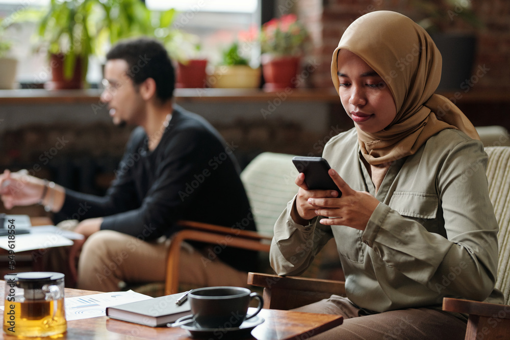 Young Muslim businesswoman in hijab messaging in smartphone or organizing work while sitting by table with tea and notebook in cafe