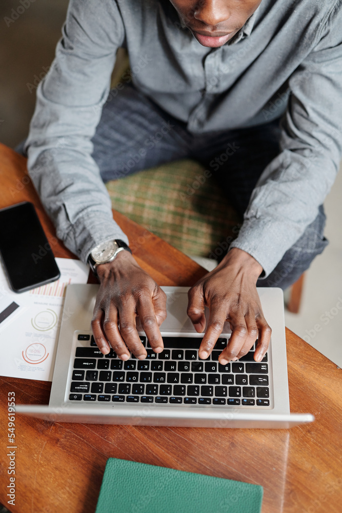 Above angle of young African American economist or analyst keeping hands over laptop keyboard while sitting by desk and organizing work Above angle of young African American economist or analyst keeping hands over laptop keyboard while sitting by desk and organizing work